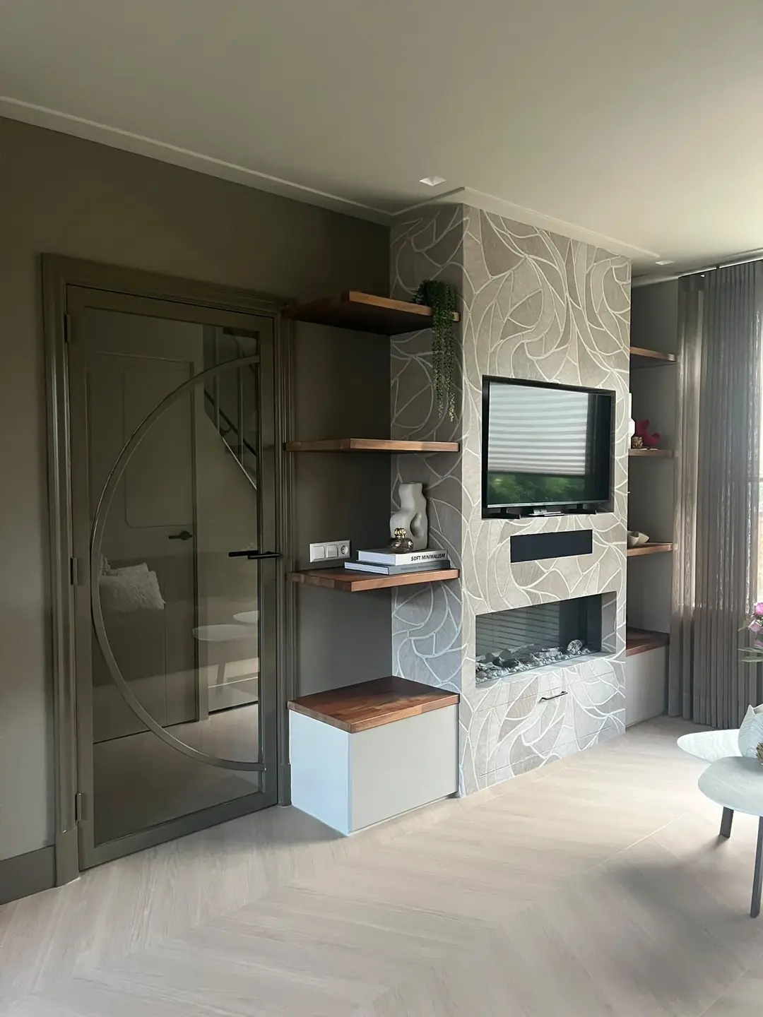 Woonkamer met tv boven de elektrische haard, wandpanelen met bladmotief behang en wandplanken van hout
