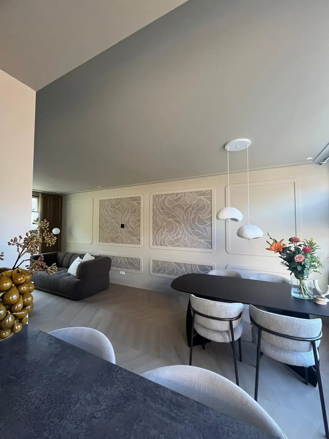 Woonkamer met wandpanelen met bladmotief behang, zwarte ovale eettafel met grijze stoelen en papieren hanglampen